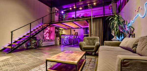 НЕОН Лофт. Лофт с антресолью, стеклянные ограждения, лестница с металлическими перилами. Диван, кресло, низкий стол. Неоновая вывеска, растения. Фиолетовая и теплая подсветка. Кирпичные стены, кафельный пол. Барная стойка с граффити. Чёрные шторы.