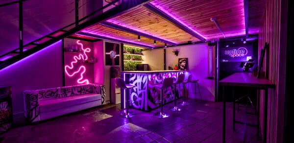 НЕОН Лофт. Современное помещение с барной стойкой, двумя барными стульями, диваном и неоновыми украшениями. Лестница на верхний уровень, металлические и деревянные отделочные материалы. Неоновая розовая подсветка, граффити на стенах и барной стойке.