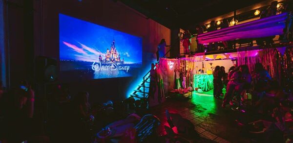 НЕОН Лофт. Темное помещение с проектором, показывающим анимацию с замком. Справа барный стол со светодиодной подсветкой и людьми, украшения с блестящими занавесками. На заднем плане подиум с прожекторами.