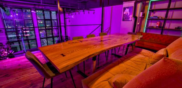 НЕОН Лофт. Современное лофт-пространство с длинным деревянным столом и стульями, угловым диваном, книжными полками. Лампы с теплым светом и фиолетовая неоновая подсветка. Большие окна и деревянный пол. На стене картина.