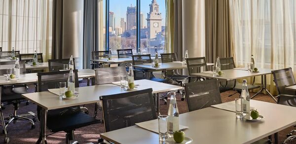 Конференц-зал Экспресс Махараджей. Конференц-зал с большими окнами и видом на город, расставлены прямоугольные столы с черными стульями на колесиках. На столах размещены бутылки воды, стаканы и зеленые яблоки. Светлые шторы, ковровое покрытие.