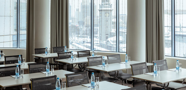 Конференц-зал Экспресс Махараджей. Конференц-зал с большими окнами, прямоугольные столы с расставленными бутылками воды и стаканами. Серые стулья на колесиках. На улице зимний пейзаж. Светлые шторы, нейтральные тона интерьера, хорошее естественное освещение.