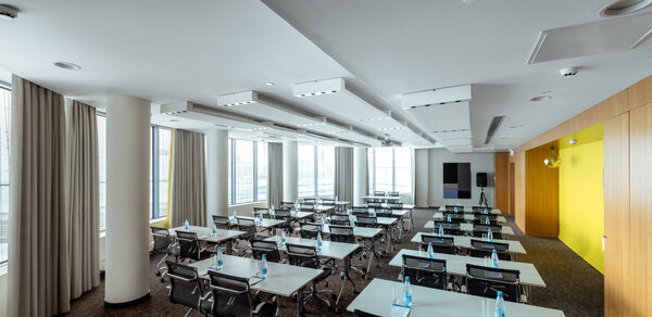 Конференц-зал Экспресс Махараджей. Конференц-зал с рядами столов, окружённых черными стульями. Ярко-желтая стена слева, окна с бежевыми шторами справа. Белый потолок с встроенным освещением. Темный ковролин на полу. Пластиковые бутылки с водой на столах.