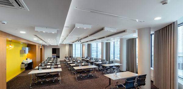 Конференц-зал Экспресс Махараджей. Конференц-зал с прямоугольной планировкой, большого размера окна. Ряд столов с черными стульями. Белые потолочные светильники, стены из светлого дерева с желтыми акцентами. Ковровое покрытие темного цвета. Шторы бежевые.