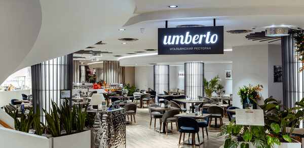 Итальянский ресторан Umberto(Умберто). Современное ресторанное пространство с круглыми столиками и мягкими стульями. Светлые деревянные полы, декоративные растения, светильники на потолке. На вывеске надпись «италиянский ресторан». Стойка из металла.