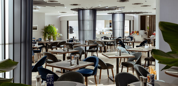 Итальянский ресторан Umberto(Умберто). Просторное помещение ресторана с расставленными квадратными столиками и темными стульями, большая барная стойка на заднем плане. Светлые стены и потолок с встраиваемыми светильниками. Декоративные растения, стеклянные и металлические элементы.