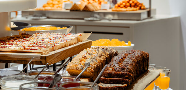 Итальянский ресторан Umberto(Умберто). Деревянные подносы с нарезанными пирогами на столе. Рядом банки с джемом и ёмкости для напитков. На заднем плане видны тарелки с пирожными и выпечкой. Пастельные оттенки, современное оформление, яркое освещение.