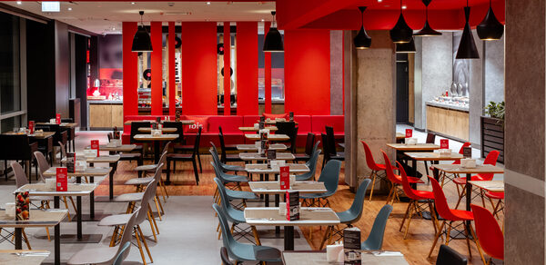 Ресторан Ibis Kitchen(Ибис Кухня). Современное кафе с открытой планировкой. Столы и стулья в нескольких рядах. Комбинация черных и серых стульев, столешницы светлого дерева. Акцентные красные детали, включая стены и стулья. Черные подвесные светильники. Пол из светлого дерева.