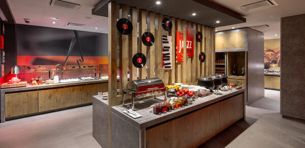 Ресторан Ibis Kitchen(Ибис Кухня). Ресторанное пространство с сервировочным островом. На столе блюда и напитки. Интерьер из дерева и камня. Настенные украшения: виниловые пластинки и постеры. Современное освещение. В тёплых тонах, доминируют коричневый и красный цвета.