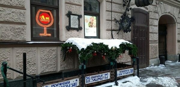 Бар "Бездна" на Маяковской. Фасад здания с декоративными элементами: светодиодный знак в виде бокала, две арочные окна, декоративная вывеска в форме бочки. Под окнами настил с зимним декором из зелени и снежным покровом, таблички с надписями рекламного характера.