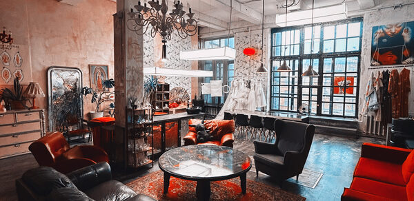 Лофт Делюкс. Лофт-пространство с высокой потолочной конструкцией и индустриальными окнами. Мебель: кожаные и мягкие кресла, круглые столы, комоды. Освещение: современные люстры и лампы. Декор на стенах, ковры на деревянном полу. Цвета: темный, красный, бежевый.