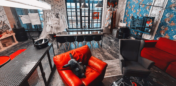 Лофт Делюкс. Лофт-пространство, декорированное в индустриальном стиле с большими окнами. В центре длинный деревянный стол с черными стульями. Красное и серое кресла, яркий диван. Декоративные элементы на стенах, большие подвесные светильники, темное напольное покрытие.