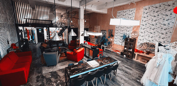 Лофт Делюкс. Лофт-пространство с бетонным потолком и колоннами, металлическими светильниками и деревянным полом. В центре длинный стол с черными стульями, кожаные кресла и красные диваны. На стенах декор и растения, повсюду яркое освещение.