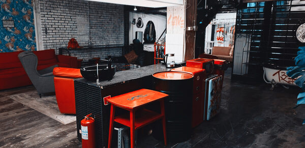 Лофт Делюкс. Лофт-пространство с барной стойкой из железных бочек и деревянным столом; кирпичные стены, деревянный пол, красная и серая мебель, большое центральное освещение; имеются диваны, кресла, раковина, огнетушитель; декорированные стены и стены с графическими рисунками.