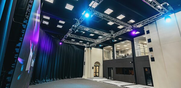 New Media (Нью Медиа) продакшн. Павильон с большим LED-экраном. Просторная комната с высокими потолками и черными занавесами. Освещение из встроенных потолочных светильников и прожекторов. Металлические конструкции для оборудования. Стены светлые, полы серые. Видны стеклянные перегородки и двери.