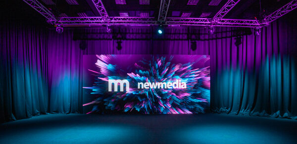 New Media (Нью Медиа) продакшн. Павильон с большим LED-экраном. Темное помещение с черными шторами и натяжным потолком с фиолетовой подсветкой. В центре светодиодный экран с цветным анимированным изображением и текстом "new media". Пол темного оттенка, система освещения закреплена на металлических конструкциях.