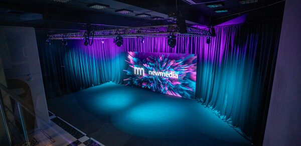New Media (Нью Медиа) продакшн. Павильон с большим LED-экраном. Темное помещение с черным потолком и оборудованием для освещения. Стены оформлены черными шторами. На полу темное покрытие. Спереди большой экран с яркими графиками и текстом "newmedia". Освещение фиолетовое и голубое.
