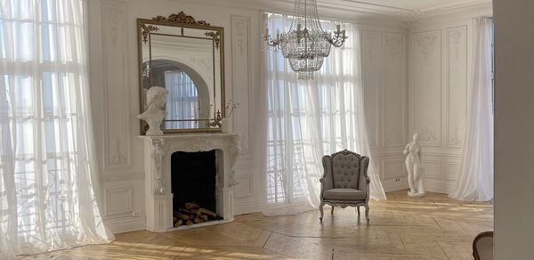 Арт-Плейс Фактура. Просторный лофт в классическом стиле. Просторное белое помещение с высоким потолком, украшенным люстрой. Паркеты из светлого дерева. Камин с декоративной рамой и скульптурами. Возле окна кресло серого цвета. Высокие окна занавешены легкими прозрачными шторами.