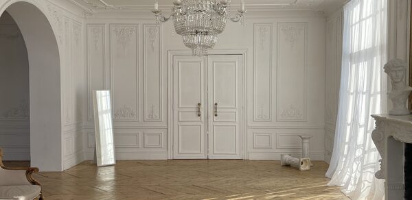 Арт-Плейс Фактура. Просторный лофт в классическом стиле. Светлая комната с паркетным полом, белыми стенами с декоративной лепниной и двойными дверями. Хрустальная люстра висит на потолке. Справа - камин и скульптура, слева - большое зеркало и арка. Под окном - занавески.