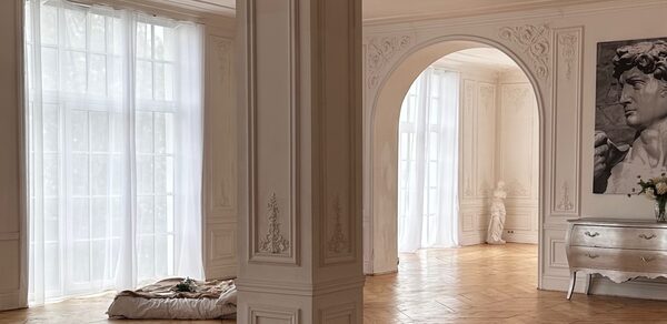 Арт-Плейс Фактура. Просторный лофт в классическом стиле. Просторное помещение с белыми резными стенами и колонной, паркетным полом, большими окнами с полупрозрачными шторами. В углу арочная ниша с белой статуей. Справа консольный стол серого цвета с декором и цветами.