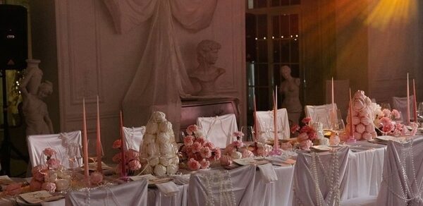 Арт-Плейс Фактура. Просторный лофт в классическом стиле. Столовая зона с длинным столом, накрытым белой скатертью и украшенным розовыми цветами и свечами. На заднем плане скульптуры и драпировки. На фоне виден мягкий свет, создающий атмосферу. Стулья обтянуты белыми чехлами.