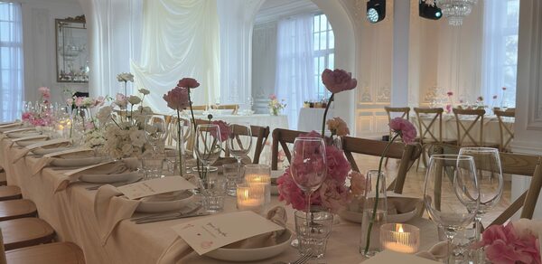 Арт-Плейс Фактура. Просторный лофт в классическом стиле. Белый банкетный зал с длинным деревянным столом, накрытым светлой скатертью. Установлены деревянные стулья. На столе стеклянная посуда, цветочные композиции, свечи. Белые стены, арочные окна, хрустальные люстры, текстильные драпировки.