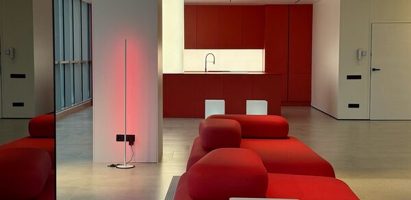 Арт-Плейс Фактура. Современный лофт с видом на весь город. Современное помещение с открытой планировкой, красные диваны, красная кухня с встроенной раковиной, барная стойка с двумя высокими стульями, светлый пол, вертикальная световая панель, большие окна слева, светлые стены.