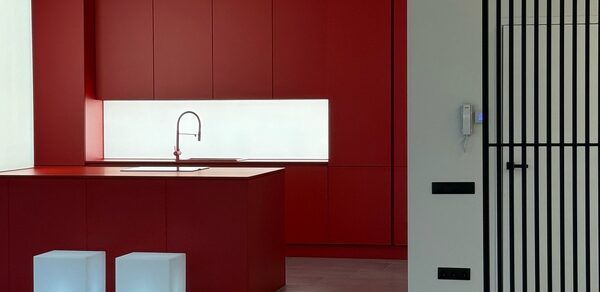 Арт-Плейс Фактура. Современный лофт с видом на весь город. Красная кухня с островом, встроенная раковина и высокий смеситель. Шкафы с закрытыми фасадами. Два белых кубических стула. Перегородка из черных вертикальных прутьев справа. Белая стена со встроенным домофоном и черными розетками. Светлые полы.