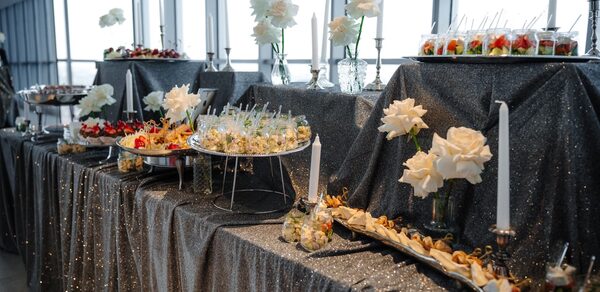 Арт-Плейс Фактура. Современный лофт с видом на весь город. Фуршетный стол с темной блестящей скатертью, сервированный закусками и десертами. Использованы возвышения для блюд. Декор из белых роз и свечей в подсвечниках. Большие окна на заднем плане обеспечивают естественное освещение.