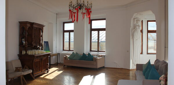 Cassiopea(Кассиопея). Просторная светлая комната с деревянным паркетом, белыми стенами и высокими потолками. Установлены два дивана и кресла с подушками. В центре — люстра с декоративными элементами. Несколько больших окон. Комод с напитками.