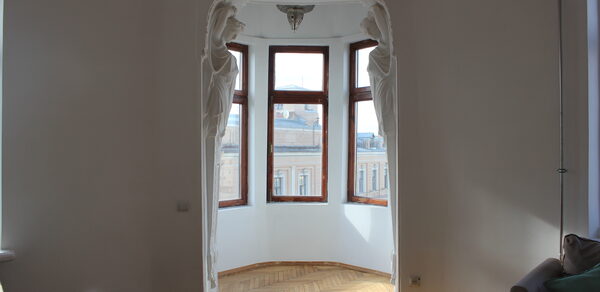 Cassiopea(Кассиопея). Эркер с большими окнами, деревянный паркет, светло-белые стены, барельефы с фигурным декором, потолочный светильник, частично виден диван с подушками.