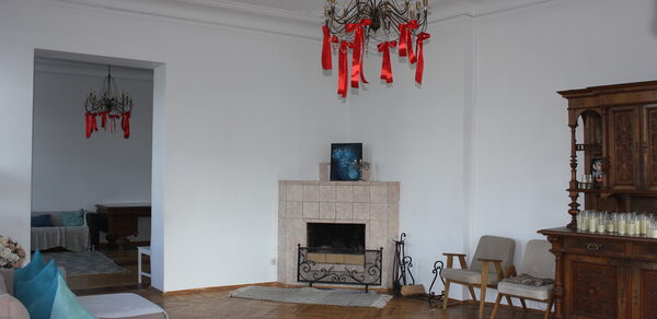 Cassiopea(Кассиопея). Гостиная комната с паркетным полом, белыми стенами и кирпичным камином. Два крупных светильника с красным декором. Мягкие диваны, деревянный шкаф с множеством свечей. Светлый интерьер, кресла и столик у стены.