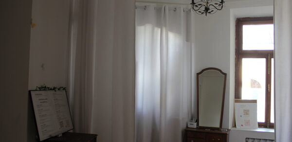 Cassiopea(Кассиопея). Комната с белыми стенами и шторами. В центре зеркало на деревянном комоде. Правее окно с белой рамой и радиатором. На комоде картины в рамках. В левом углу стоит стол с черной доской. Потолок оформлен черной люстрой.