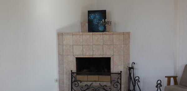 Cassiopea(Кассиопея). Камин из светлой плитки с кованой решеткой, рядом каминные принадлежности. На камине декоративная картина и статуэтка. В углу кресло, на полу ковер. Светлые стены и паркетный пол.