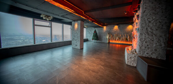 ATMOSFERA 53(Атмосфера 53). Просторное помещение с большими окнами и видом на город, темный пол и потолок, декоративные колонны и стены с текстурой камня, елка и растительные композиции, мягкое освещение с теплым оттенком.