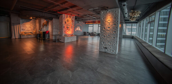 ATMOSFERA 53(Атмосфера 53). Просторное помещение с бетонными колоннами и стенами, отделанными декоративной штукатуркой. Пол выложен темным кафелем, множество больших окон с видом на город. Имеется зона с креслами, декоративный камин. Освещение теплое, с акцентом на колонны.