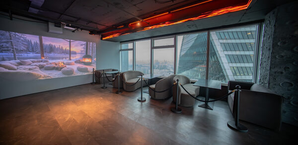 ATMOSFERA 53(Атмосфера 53). Современное помещение с панорамными окнами и видом на город. Серые кресла и столы расположены вдоль окон. На полу плитка темного цвета. Потолок с открытыми конструкциями и встроенным освещением. На стене проекция зимнего пейзажа.