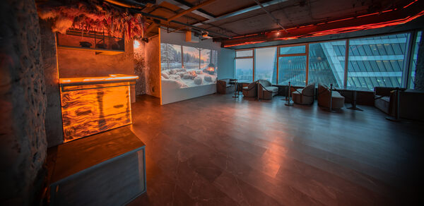 ATMOSFERA 53(Атмосфера 53). Просторное помещение с панорамными окнами и видом на город. Лофт-стиль, подвесной потолок с открытыми коммуникациями, естественное и неоновое освещение. Барная стойка из дерева, зона с мягкими креслами и столами у окон. Темный пол из плитки.