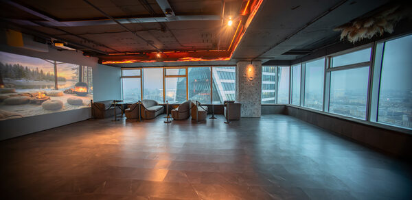 ATMOSFERA 53(Атмосфера 53). Просторное помещение с большими окнами, оклеенными изнутри жалюзи, вид на городские здания. Полы из темного материала, потолок с трассированными светильниками. Несколько стульев и столики у панорамных окон. На стене экран с изображением.