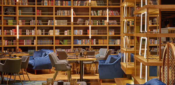 Communitymoscow (Коммьюнити Москоу).Зал Библиотека. Многофкнкциональное пространство. Библиотечное пространство с высокими деревянными полками, заполненными книгами. Мягкие кресла серого и синего цвета вокруг круглых столов. Теплое освещение с вертикальными световыми полосами. На столах стеклянные бокалы янтарного цвета.