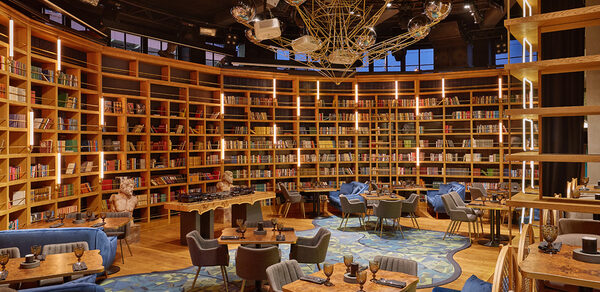 Communitymoscow (Коммьюнити Москоу).Зал Библиотека. Многофкнкциональное пространство. Библиотека с высокими полками, полными книг, зона отдыха с креслами и столиками. Деревянная мебель, мягкие серые кресла, синий ковер. Потолочные светильники и металлическая люстра. Теплое освещение, деревянный пол.