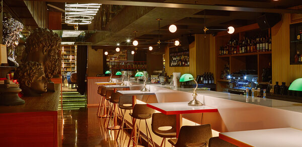 Communitymoscow (Коммьюнити Москоу).Зал Библиотека. Многофкнкциональное пространство. Барное пространство с длинными столами и стульями, на столах зеленые лампы. Потолочные светильники с круглыми плафонами, задняя стенка со стеллажами для бутылок. Интерьер в теплых тонах, темный пол, зеркальные элементы. Подвесной неоновый знак.