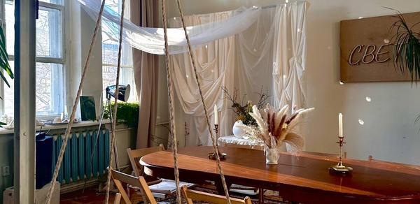 Студия Свети. Просторная комната с деревянным столом и складными стульями. На столе свечи и композиция из сухих трав. Стена декорирована светлой тканью, на стенде надпись кириллицей. Окна с прозрачными и светлыми шторами, зелёные радиаторы.