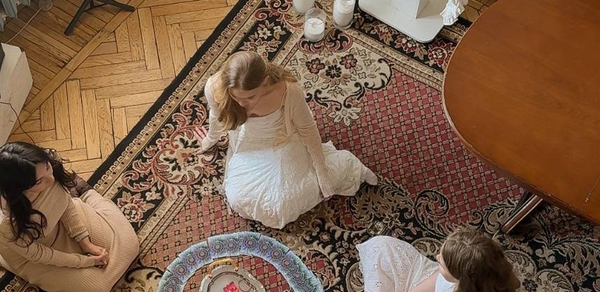 Студия Свети. Гостиная с паркетным полом и ковром с узорами, овальный деревянный стол, вокруг которого сидят три женщины в светлых платьях. На полу круглый поднос со свечами и цветком в воде, рядом горят свечи в стеклянных подсвечниках.