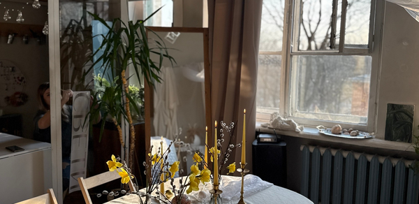 Студия Свети. Комната с большим окном и радиатором, круглый стол с белой скатертью, украшенный цветами и свечами. Деревянные стулья, зеркало, комнатное растение. Светлые шторы, на подоконнике декор, отражение человека в зеркале. Естественное освещение.