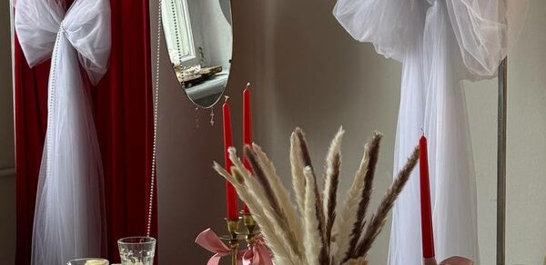 Студия Свети. Стол с белой скатертью, украшенный букеты и красными свечами на золотистых подсвечниках. Справа зеркало в круглой раме, на фоне красные шторы с белым текстильным бантом. Бежевые стены, естественное освещение.