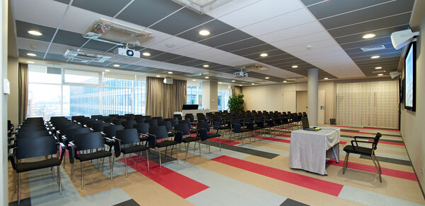 Конференц-зал ТехноПрогресс. Конференц-зал с рядами черных стульев, проекторы на потолке, окно с плотными шторами, стол с белой скатертью, разноцветное ковровое покрытие в серых и красных тонах, светло-серые стены, встроенные светильники.