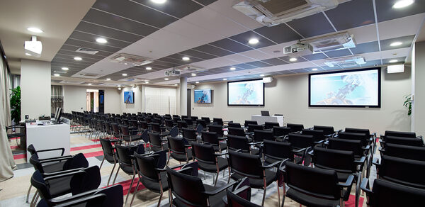 Конференц-зал ТехноПрогресс. Конференц-зал с рядами черных стульев, три экрана на стене, место для выступающего с белым пюпитром. Потолок с встраиваемыми светильниками и вентиляционными системами. Светло-серое и красное напольное покрытие.