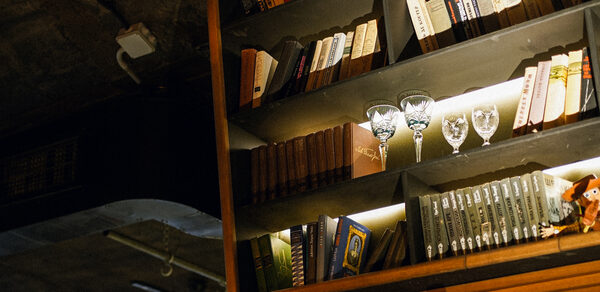 Гастробар Дневник. Книжная полка с разноцветными книгами и бокалами для вина, освещенная встроенной подсветкой. Потолок из темного бетона, видны элементы освещения и вентиляции. Доминируют теплые тона.