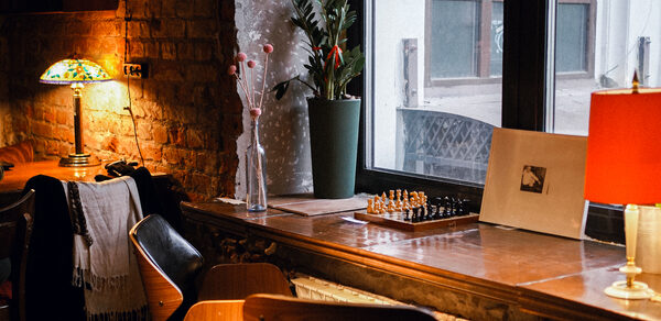 Гастробар Дневник. Лофт-пространство с кирпичными стенами и деревянной мебелью. На стене витражная лампа, настольная игра, вазоны с растениями. Красная настольная лампа и кресла. Большое окно с видом на соседнее здание.